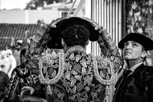 El peso de la tarde - La mirada del subalterno. Fotografía de Javier Pérez mostrando a un matador de espaldas ajustándose la montera frente a un hombre de plata.